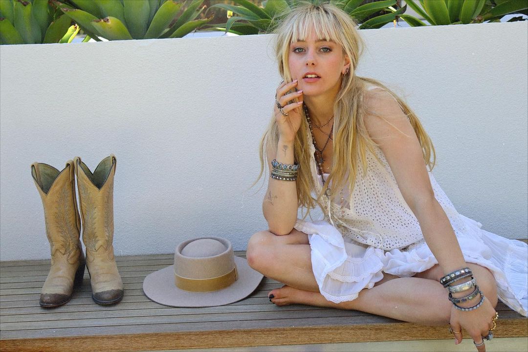 Ellery Sprayberry sitting on a bench with boots and a hat beside her