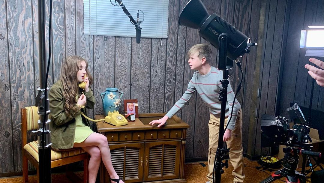 Paxton Mishkind leaning on a cabinet. A girl is talking on a phone sitting in a chair.
