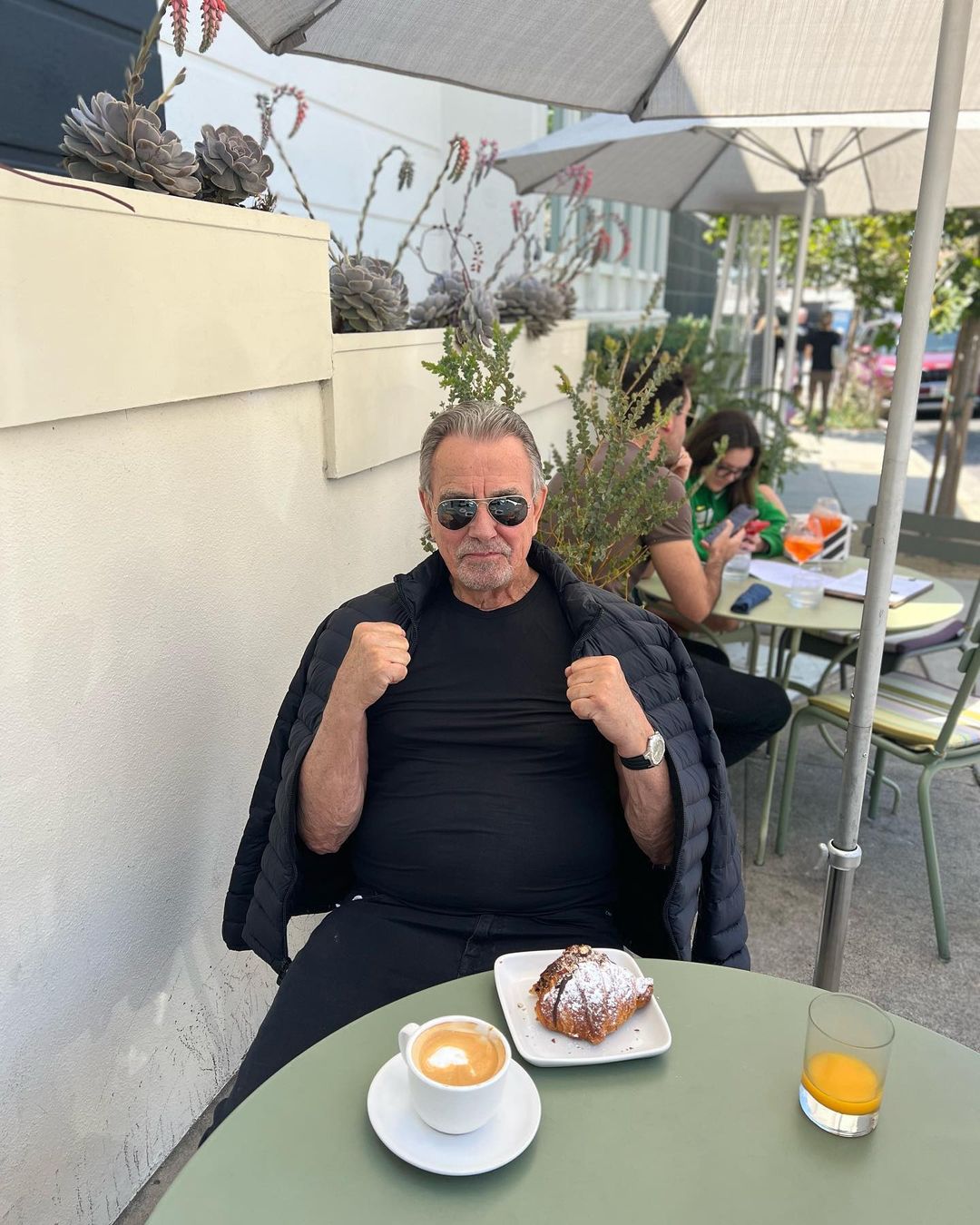 Eric Braeden enjoying his meal and giving a pose to the camera with his fist closed and black puffy jacket over his shoulder.