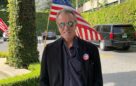 Eric Braeden standing in front of the flag of USA with I Voted badge on his jacket.