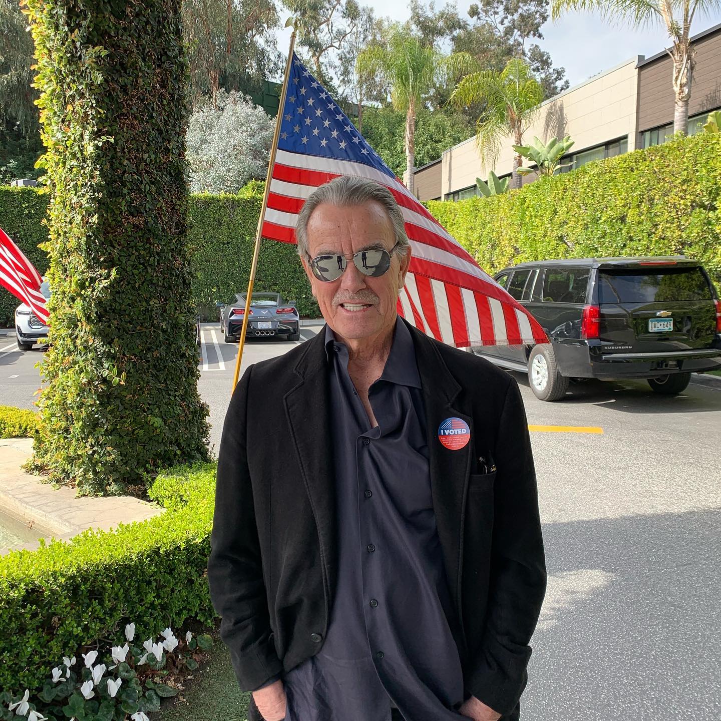 Eric Braeden standing in front of the flag of USA with I Voted badge on his jacket.