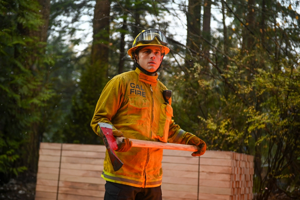 Zach Tinker wearing firefighter gear and holding an axe in his hand as he shoots for Fire Country.